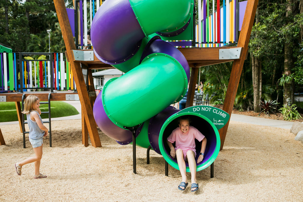 A girl wearing a pink shirt exits a spiral playground slide while another girl walks nearby on the playground's sandy surface.