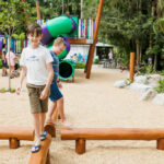 Two boys balance on a wooden beam at an outdoor playground with slides, climbing structures, and trees in the background.