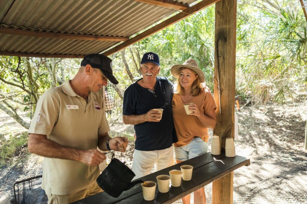 Three adults outdoors at a wooden counter; one man pours drinks into paper cups while another man and a woman stand together holding cups, surrounded by greenery.