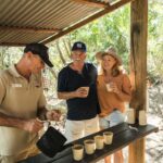 Three adults outdoors at a wooden counter; one man pours drinks into paper cups while another man and a woman stand together holding cups, surrounded by greenery.