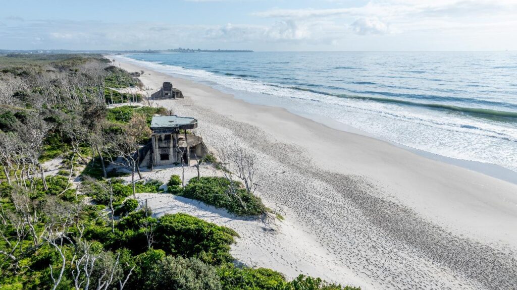 Aerial view of a sandy beach with ocean waves, dense coastal vegetation, and a concrete bunker structure near the shoreline.