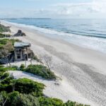 Aerial view of a sandy beach with ocean waves, dense coastal vegetation, and a concrete bunker structure near the shoreline.
