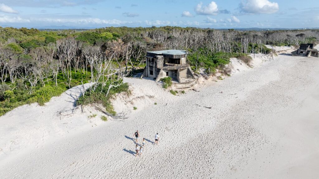Aerial view of a sandy beach with three people walking near old concrete bunkers, bordered by dense coastal vegetation.