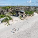 Aerial view of a sandy beach with three people walking near old concrete bunkers, bordered by dense coastal vegetation.
