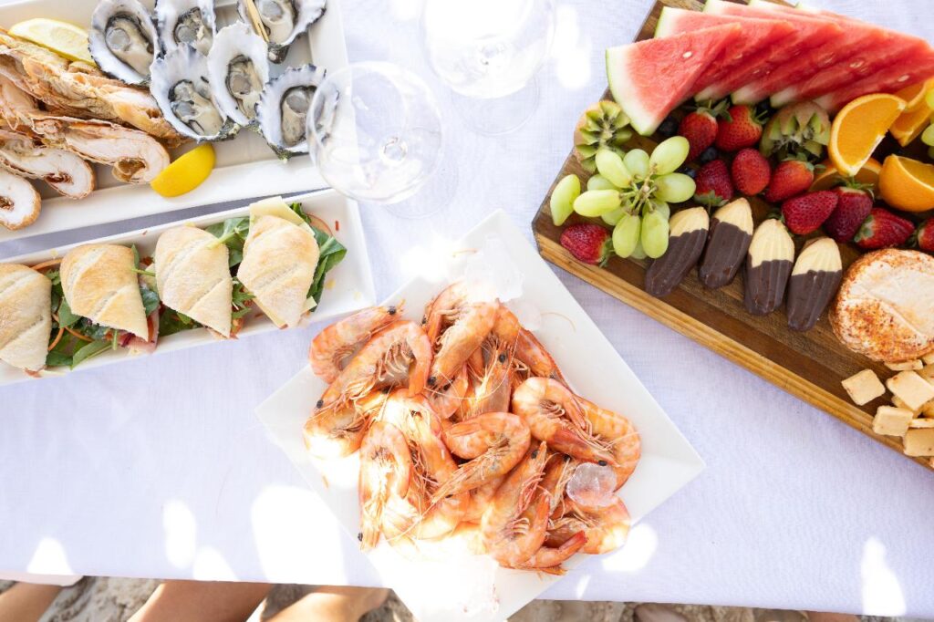 A table set with plates of shrimp, oysters, sandwiches, and a fruit platter with strawberries, grapes, watermelon, and orange slices on a white tablecloth.