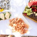 A table set with plates of shrimp, oysters, sandwiches, and a fruit platter with strawberries, grapes, watermelon, and orange slices on a white tablecloth.