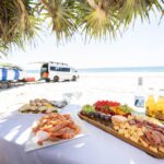 A table on a sandy beach displays seafood, cheese, fruit, and drinks. In the background, a van and surfboards are parked near the ocean under a sunny sky.