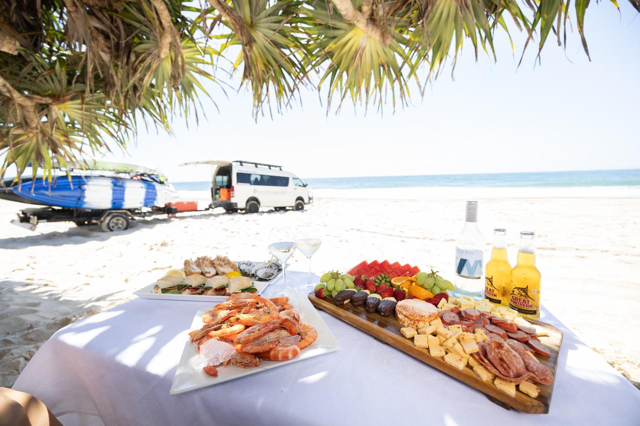 A table on a sandy beach displays seafood, cheese, fruit, and drinks. In the background, a van and surfboards are parked near the ocean under a sunny sky.