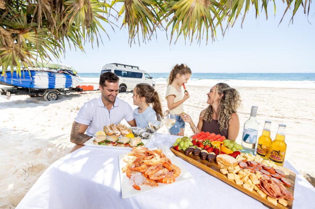 A family of four enjoys a seafood and fruit picnic at a table on a sandy beach with a van and surfboards in the background.