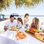 A family of four enjoys a seafood and fruit picnic at a table on a sandy beach with a van and surfboards in the background.