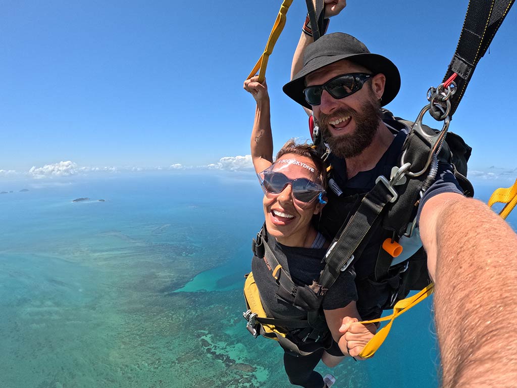 Two people tandem skydiving over a clear blue ocean with coral reefs and islands visible below, both smiling at the camera mid-air.