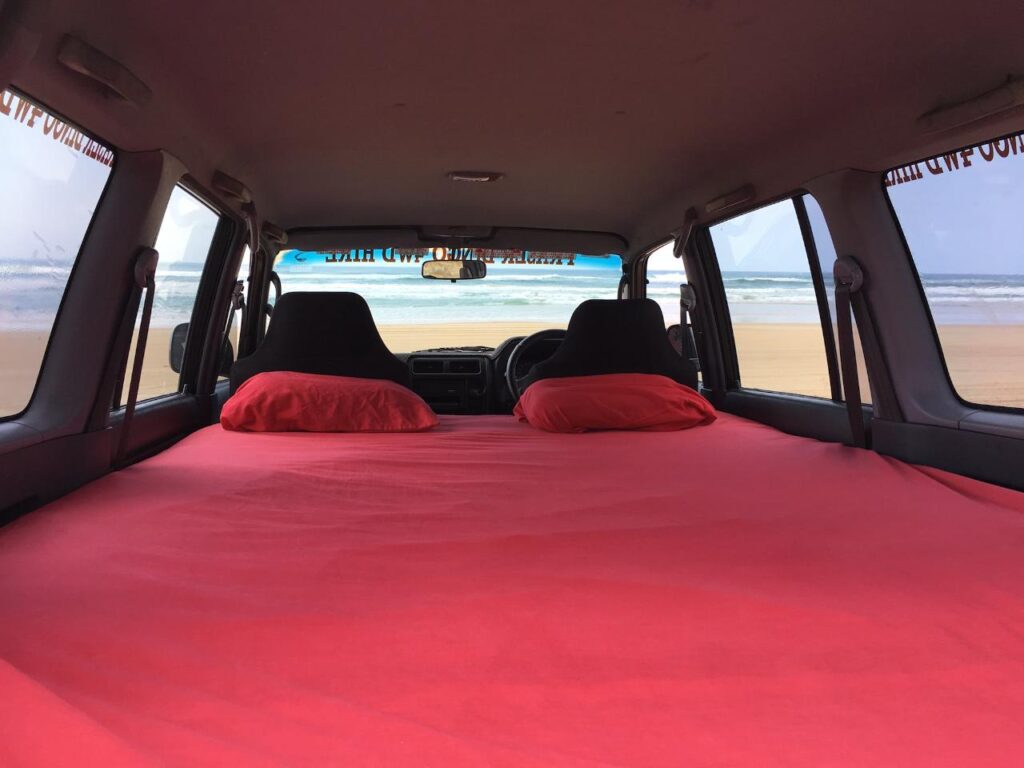 View from inside a vehicle with a red-sheeted bed setup, facing forward towards the beach and ocean visible through the windshield.