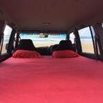 View from inside a vehicle with a red-sheeted bed setup, facing forward towards the beach and ocean visible through the windshield.