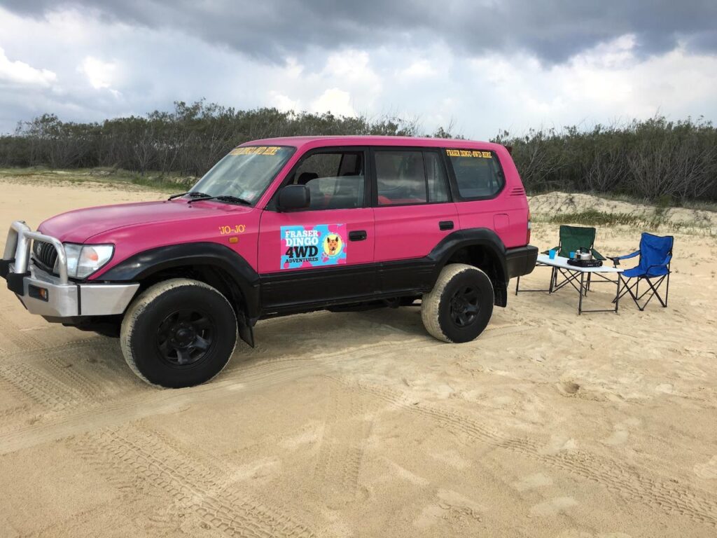 A pink 4WD vehicle parked on sand near a foldable table with camping gear and two blue chairs; bushland and cloudy sky in the background.