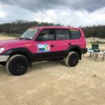 A pink 4WD vehicle parked on sand near a foldable table with camping gear and two blue chairs; bushland and cloudy sky in the background.