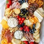 Charcuterie board with assorted meats, cheeses, crackers, fresh fruits, nuts, dips, and bread slices arranged on a wooden board.
