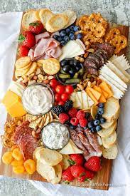 Charcuterie board with assorted meats, cheeses, crackers, fresh fruits, nuts, dips, and bread slices arranged on a wooden board.