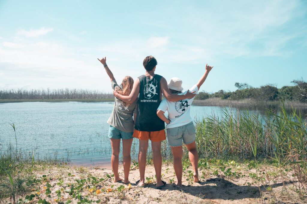 Three people stand on a sandy bank facing a body of water, arms raised and linked, with grassy plants and trees in the background under a clear sky.