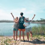 Three people stand on a sandy bank facing a body of water, arms raised and linked, with grassy plants and trees in the background under a clear sky.