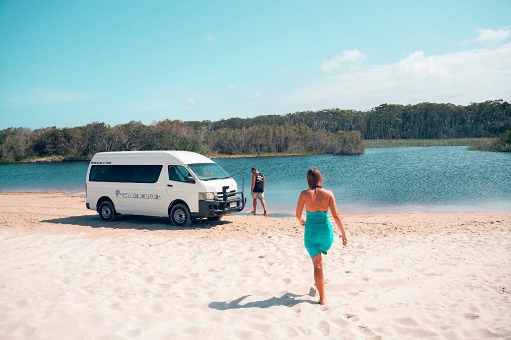 A woman walks on sandy beach toward a parked white van and another person standing near a lake with trees in the background.