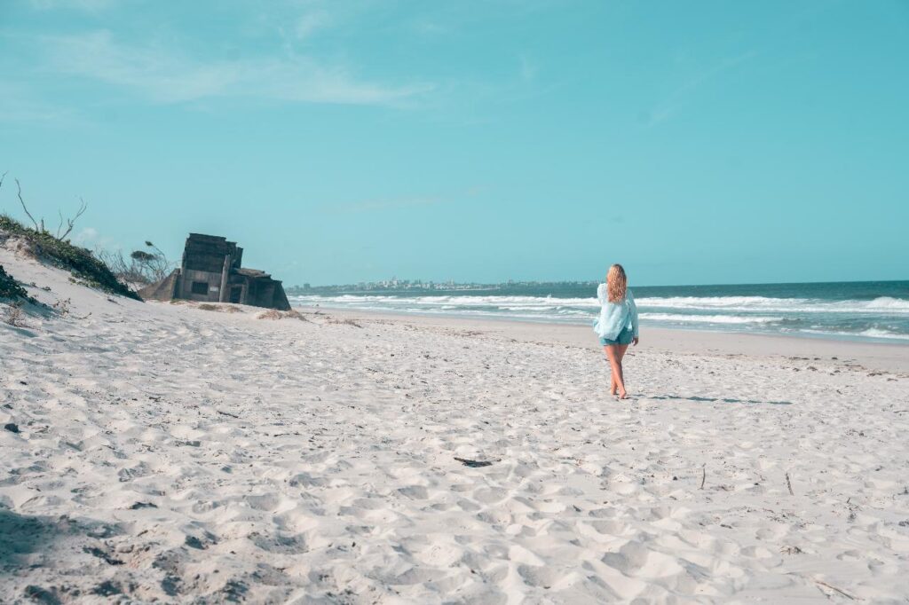 A person with long blonde hair walks alone on a sandy beach toward the ocean, with an old concrete structure visible in the distance under a clear blue sky.