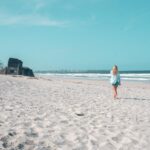A person with long blonde hair walks alone on a sandy beach toward the ocean, with an old concrete structure visible in the distance under a clear blue sky.