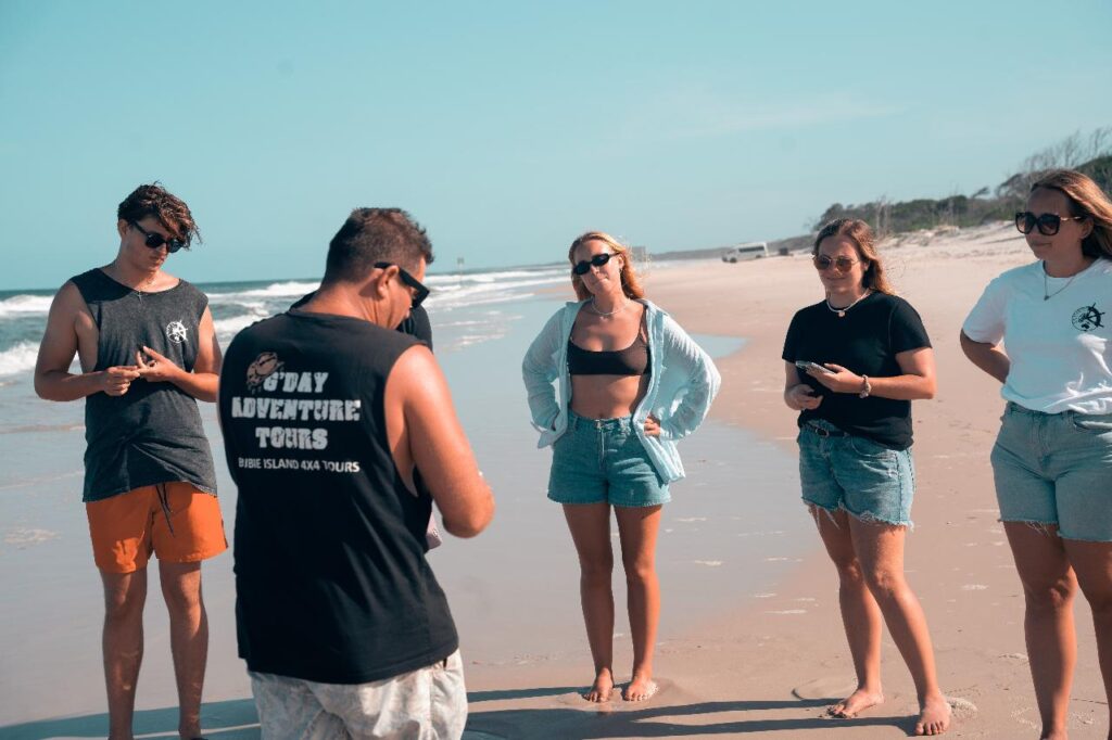 Five people stand on a sandy beach, some looking at their phones and others talking. One person wears a tank top that reads "Sav Adventure Tours." The ocean is visible in the background.