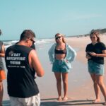 Five people stand on a sandy beach, some looking at their phones and others talking. One person wears a tank top that reads "Sav Adventure Tours." The ocean is visible in the background.