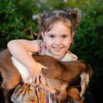 Young girl with pigtails wearing a floral dress sits on a bench, smiling at the camera while holding a brown and white baby goat outside.