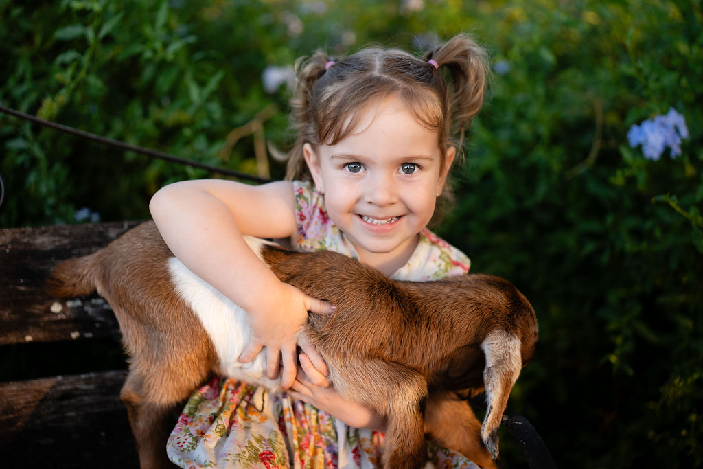 Young girl with pigtails wearing a floral dress sits on a bench, smiling at the camera while holding a brown and white baby goat outside.