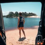 A person in shorts and a sleeveless shirt stands on a sandy shore looking at a river, viewed from inside a vehicle with equipment visible in the foreground.
