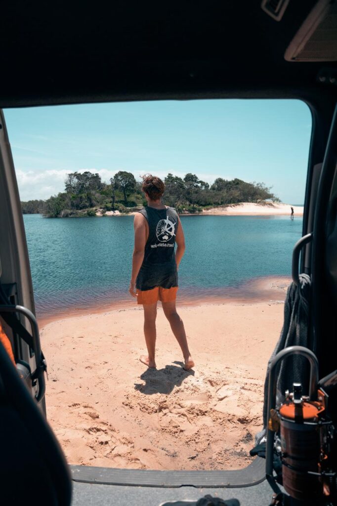 A person in shorts and a sleeveless shirt stands on a sandy shore looking at a river, viewed from inside a vehicle with equipment visible in the foreground.