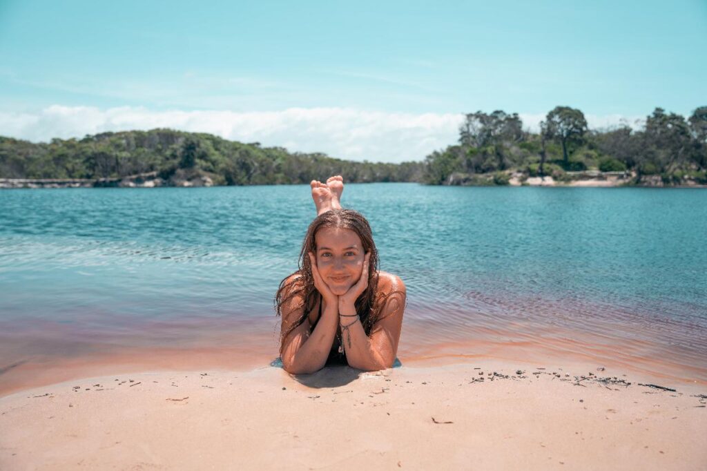 A person lies on their stomach at the edge of a lake, resting their chin in their hands, with trees and blue sky in the background.