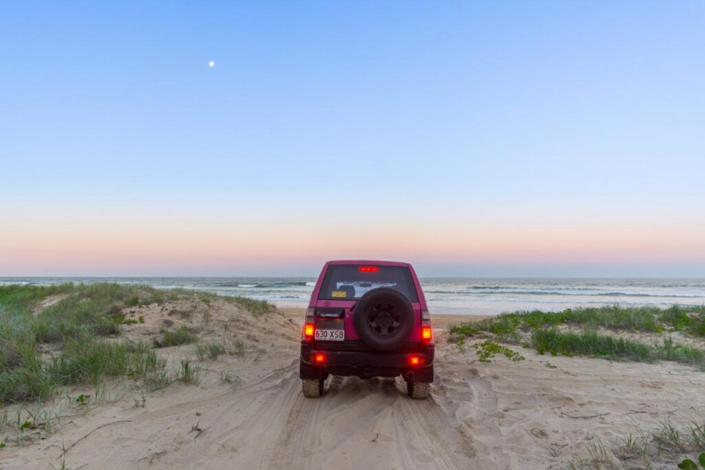 A red SUV drives on sandy terrain toward the ocean at dusk, with grass on both sides and a clear sky above.