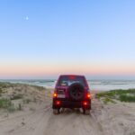 A red SUV drives on sandy terrain toward the ocean at dusk, with grass on both sides and a clear sky above.
