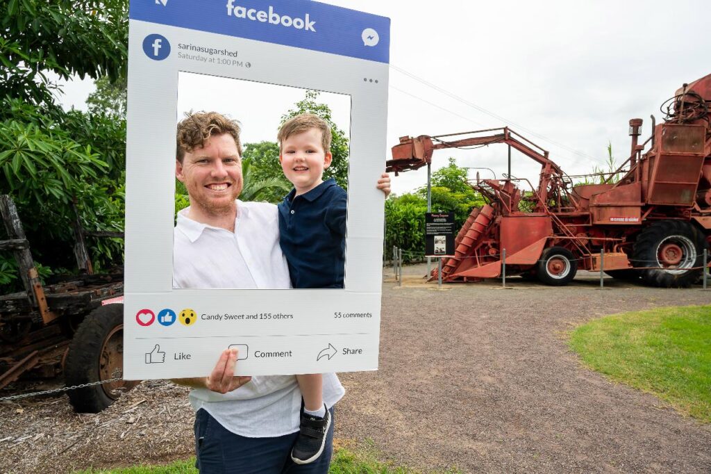 A man holds a child and a large cardboard Facebook frame prop, standing outdoors near old red farming machinery and greenery.