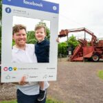 A man holds a child and a large cardboard Facebook frame prop, standing outdoors near old red farming machinery and greenery.