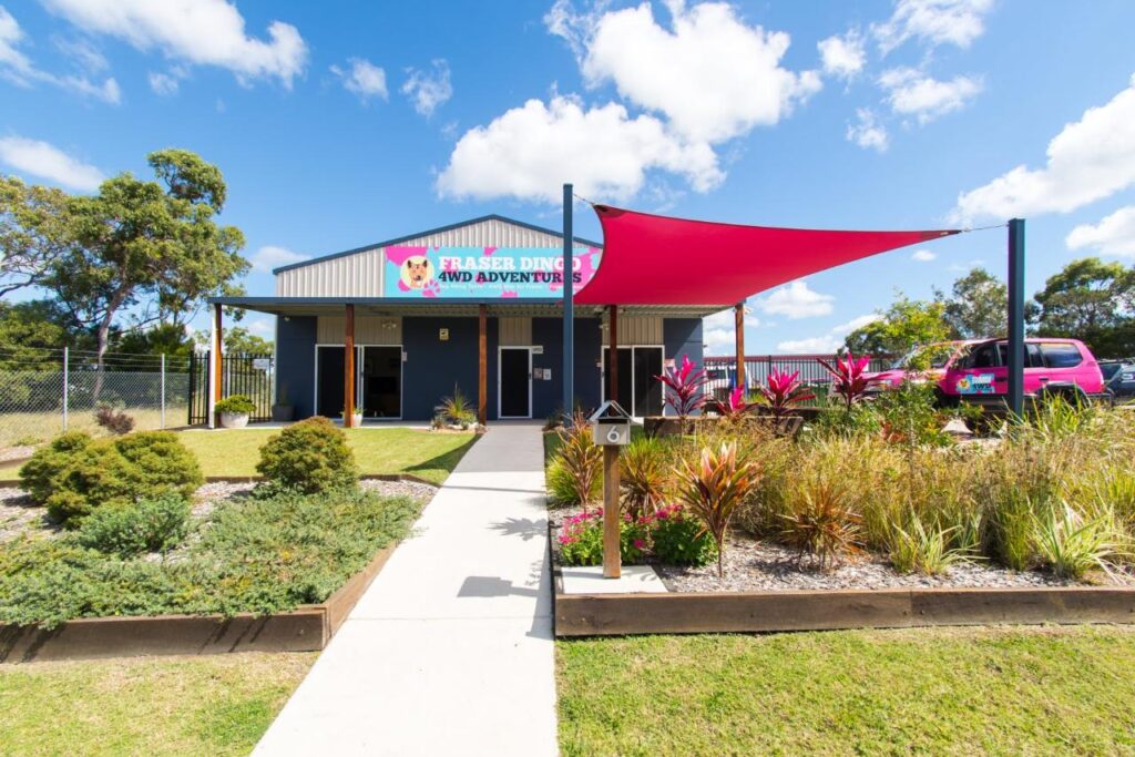 A small building with a sign reading "Fraser Ding Kidz Adventures," landscaped garden, red shade sail, and a pink vehicle parked to the right under a blue sky with clouds.