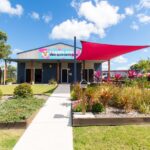 A small building with a sign reading "Fraser Ding Kidz Adventures," landscaped garden, red shade sail, and a pink vehicle parked to the right under a blue sky with clouds.