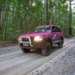 A pink 4x4 SUV drives on a sandy dirt road through a dense, green forest.