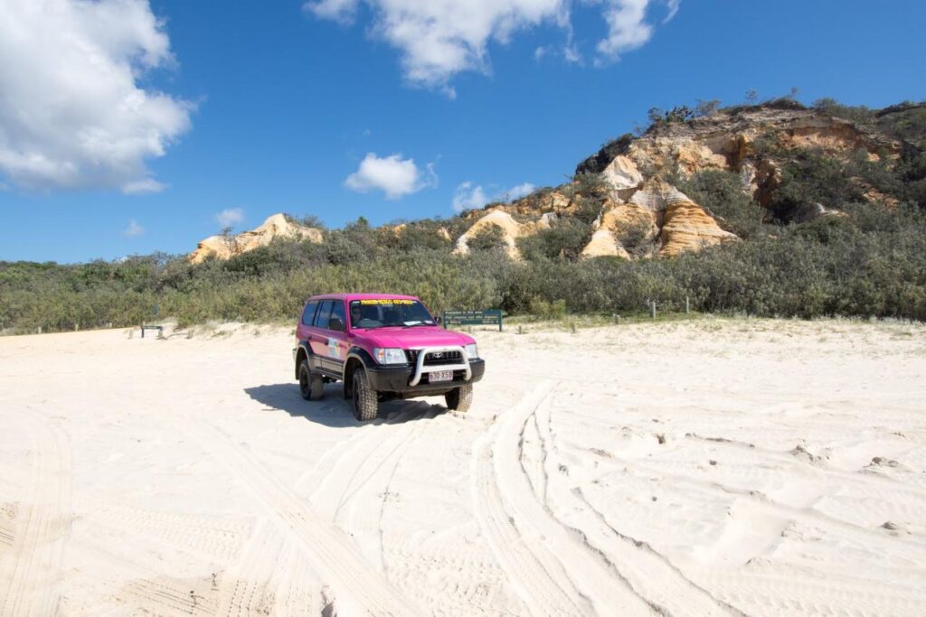 A pink SUV is parked on a sandy beach with tire tracks, shrubbery, and rocky cliffs in the background under a partly cloudy sky.