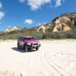 A pink SUV is parked on a sandy beach with tire tracks, shrubbery, and rocky cliffs in the background under a partly cloudy sky.