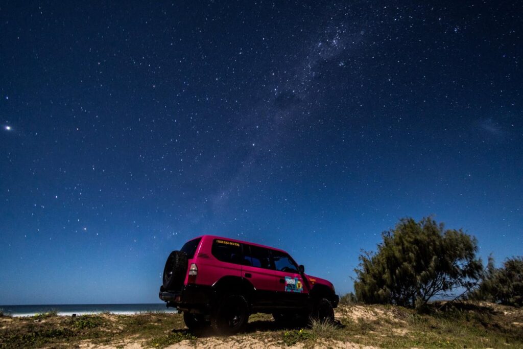 A red SUV parked on sandy ground by the ocean at night, with trees nearby and a clear sky filled with stars above.