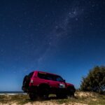 A red SUV parked on sandy ground by the ocean at night, with trees nearby and a clear sky filled with stars above.