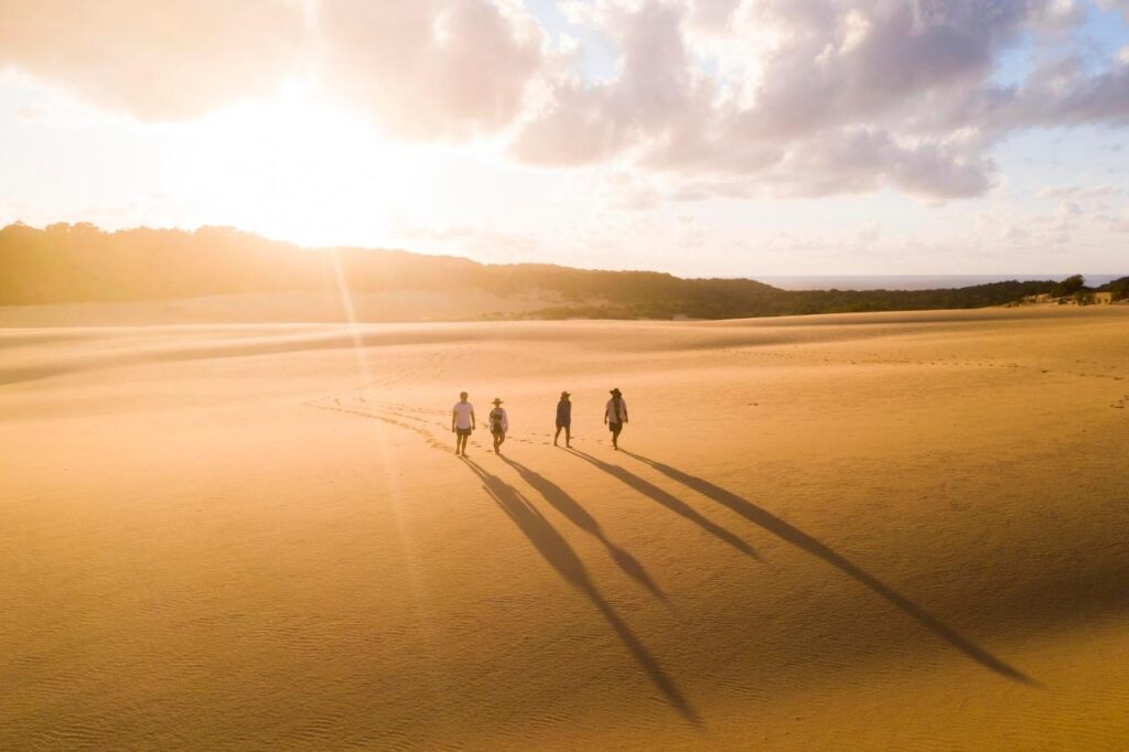 Four people walk across a sandy desert landscape at sunset, casting long shadows behind them under a partly cloudy sky.