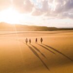 Four people walk across a sandy desert landscape at sunset, casting long shadows behind them under a partly cloudy sky.