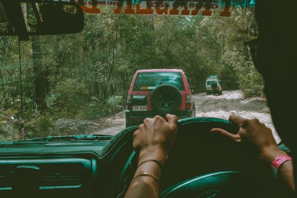 View from inside a vehicle showing hands on the steering wheel, following two cars on a dirt road surrounded by trees.