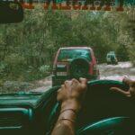 View from inside a vehicle showing hands on the steering wheel, following two cars on a dirt road surrounded by trees.