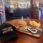 A cheeseburger with lettuce and tomato, fries in a metal basket, and a cup of sauce on a wooden board next to a black cap, set on a table in a bar-style restaurant.