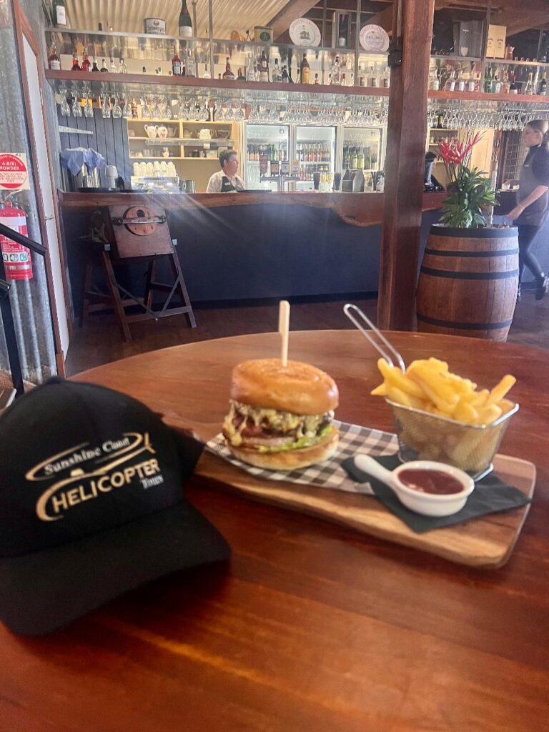 A cheeseburger with lettuce and tomato, fries in a metal basket, and a cup of sauce on a wooden board next to a black cap, set on a table in a bar-style restaurant.